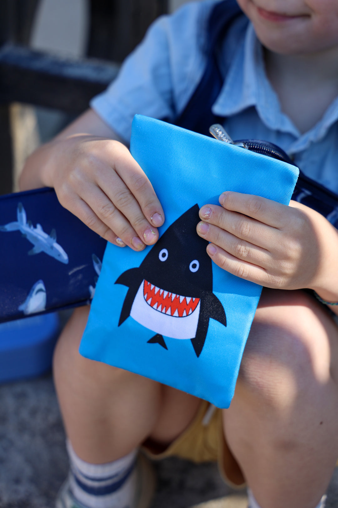 Child holding a blue pouch with a shark design, sitting outdoors.