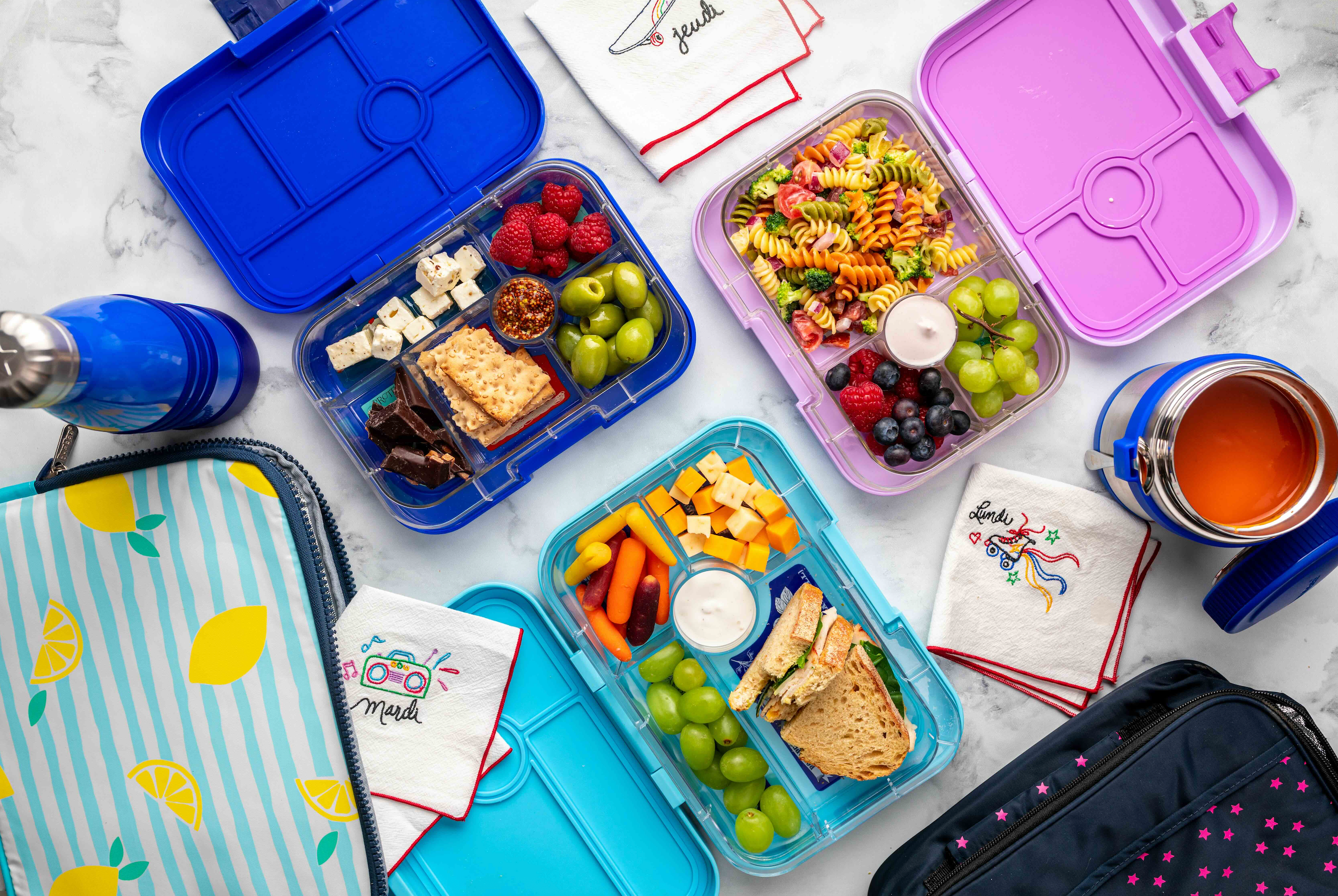 Assorted Yumbox lunch containers with food and a thermos on a marble surface